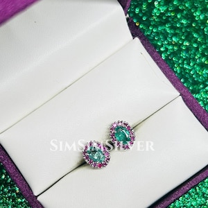 May include: A pair of oval stud earrings. Each earring has a central green gemstone encircled by smaller pink gemstones. The earrings are presented in a white jewellery box with a purple interior.