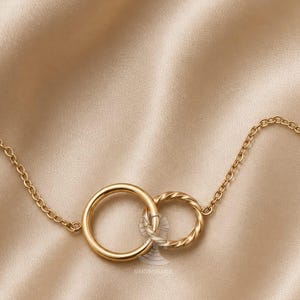 May include: A gold-colored necklace featuring two interlocking circles, one smooth and one twisted, connected by a delicate chain. The jewelry is displayed on a satin-like, beige background, creating a luxurious aesthetic.