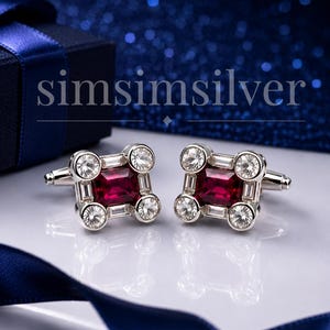 May include: A pair of silver-toned cufflinks featuring a square-cut deep red center stone surrounded by four round clear stones at the corners and small rectangular clear stones along the edges. The text simsimsilver is visible above the cufflinks.