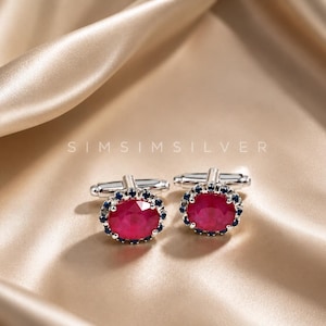 Può includere: Un paio di gemelli in argento con pietre centrali rosse di forma ovale circondate da un alone di piccole pietre blu scuro. Il testo SIMSIMSILVER è visibile sopra. Il design include un meccanismo di chiusura a levetta standard.
