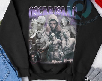 Może przedstawiać: Czarna bluza z nadrukiem graficznym przedstawiającym Oscara Isaaca jako Moon Knighta. Nadruk zawiera kolaż różnych postaci z serialu, w tym Moon Knighta, Mr. Knighta i egipskiego boga Khonshu. Tekst "Oscar Isaac" jest nadrukowany czcionką w stylu retro.