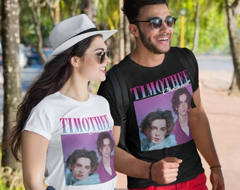 May include: Two people wearing t-shirts with a graphic of Timothée Chalamet. The shirts are black and white and feature a pink and purple background. The people are walking along a pavement.