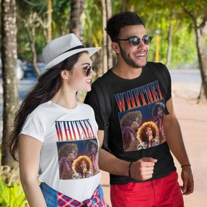 May include: Two people wearing white t-shirts with a vintage image of Whitney Houston on the front. The image is a collage of three photos of Whitney Houston performing on stage. The text "WHITNEY" is printed above the image. The people are walking down a pavement.