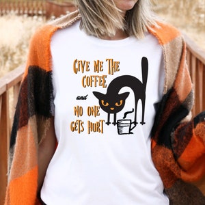 Puede incluir: Camiseta blanca con un gato negro sosteniendo una taza de café y el texto "Dame el café y nadie sale herido".