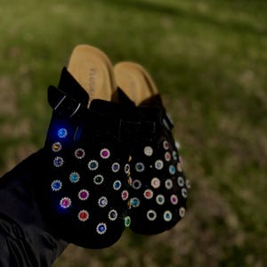 May include: Black clogs adorned with colorful rhinestones. The clogs have a suede-like texture and feature a buckle closure. The footbed is a light tan color. The clogs are held up against a blurred green background.