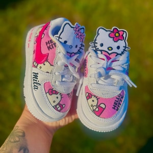 Puede incluir: Zapatillas Hello Kitty blancas y rosas. Los zapatos presentan el personaje de Hello Kitty en los laterales y la lengüeta, con el nombre "mila" escrito en el lateral. El logo de Nike es rosa.