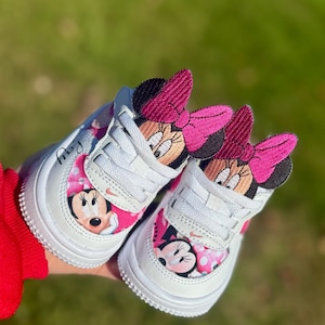 Puede incluir: Zapatillas blancas para bebé con diseños de Minnie Mouse. Los zapatos tienen lazos y detalles en rosa, con la cara de Minnie Mouse en los laterales. Los cordones son blancos y las suelas son blancas. Los zapatos se muestran sobre un fondo verde.