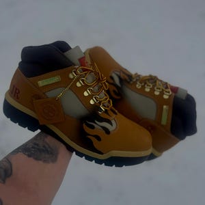 Puede incluir: Un par de botas Timberland marrones y marrones con ojales y cordones dorados. Las botas presentan un diseño de llama en el lateral y las letras "JR". Las botas tienen una suela negra y un cuello marrón.