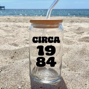 Peut inclure: Une canette en verre transparent avec un couvercle en bois brun et une paille noire. La canette porte une étiquette noire avec le texte "CIRCA 1984".