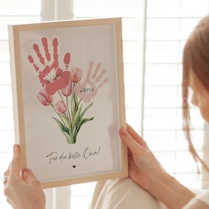 May include: A framed print with pink handprints and the names Liam and Lena. The handprints are surrounded by pink tulips and the text "F&uuml;r die beste Oma!" (For the best Grandma!).