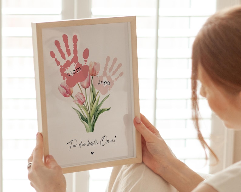 May include: A framed print with pink handprints and the names Liam and Lena. The handprints are surrounded by pink tulips and the text "F&uuml;r die beste Oma!" (For the best Grandma!).
