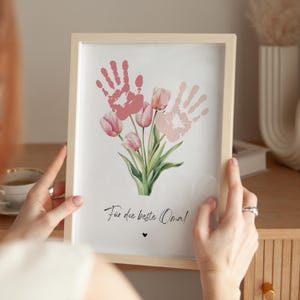 May include: A framed print with a handprint design of pink tulips. The print says "F&uuml;r die beste Oma!" which translates to "For the best Grandma!" in English.