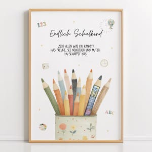 May include: A framed print with a light wood frame. The print features a watercolor illustration of a pencil holder filled with colorful pencils. Text in German reads "Endlich Schulkind" and other phrases. Decorative elements include numbers, a globe, and a clock.
