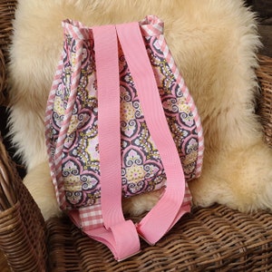 Puede incluir: Una mochila rosa y estampada con correas ajustables. La mochila presenta un dise&ntilde;o floral en rosa, amarillo y gris, con un ribete de cuadros rosa y blanco. Las correas son de color rosa liso.