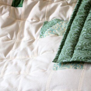 May include: Close-up of a handmade quilt featuring a patchwork design. The quilt showcases a combination of off-white and green fabrics. The green fabric has a swirling pattern, while the off-white fabric is quilted in a grid pattern.