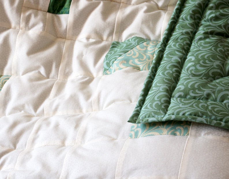 May include: Close-up of a handmade quilt featuring a patchwork design. The quilt showcases a combination of off-white and green fabrics. The green fabric has a swirling pattern, while the off-white fabric is quilted in a grid pattern.