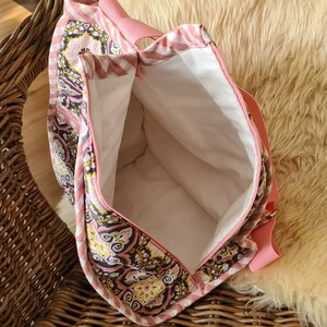 Puede incluir: Vista abierta de un bolso tote rosa y estampado. El bolso presenta un dise&ntilde;o floral y paisley en tonos rosa, negro y amarillo. El interior es blanco y el bolso tiene una cremallera y una correa rosas. El bolso est&aacute; abierto, revelando el espacioso interior.