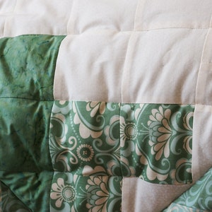 May include: Close-up of a quilted blanket with a patchwork design. The quilt features alternating squares of white fabric and green patterned fabric. The green fabric has a marbled and floral design.