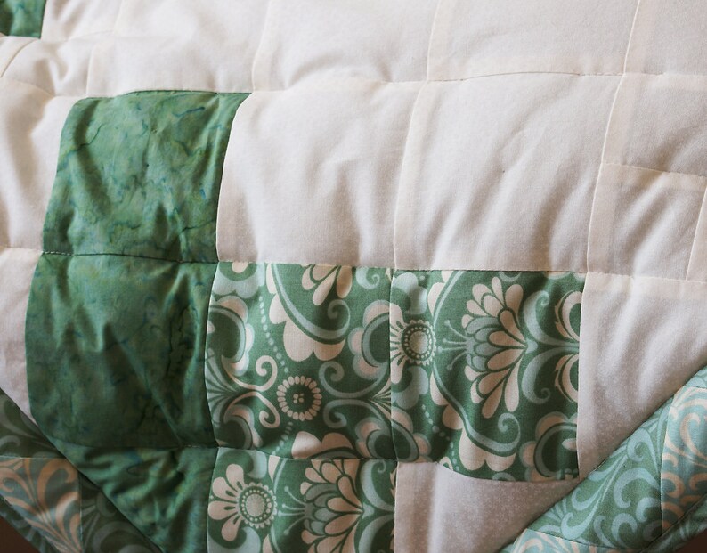 May include: Close-up of a quilted blanket with a patchwork design. The quilt features alternating squares of white fabric and green patterned fabric. The green fabric has a marbled and floral design.