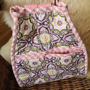 Puede incluir: Bolso tote hecho a mano con un estampado floral en rosa, gris y amarillo. El bolso presenta un ribete de cuadros rosas y blancos y un bolsillo frontal con cremallera. El asa es rosa.