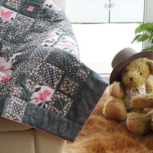 May include: A handmade patchwork quilt with a bird and floral design. The quilt features squares of gray, white, and pink fabric. A teddy bear wearing a hat sits nearby. The quilt is draped over a white surface.