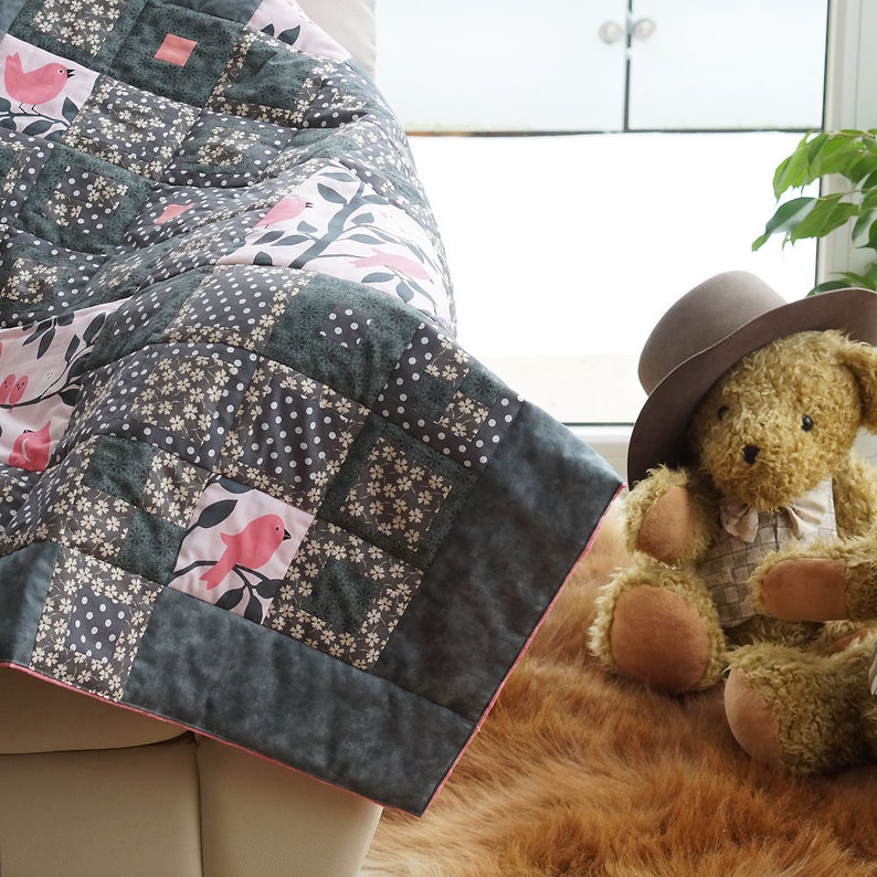 May include: A handmade patchwork quilt with a bird and floral design. The quilt features squares of gray, white, and pink fabric. A teddy bear wearing a hat sits nearby. The quilt is draped over a white surface.
