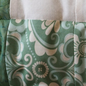 May include: Close-up of a quilt with a floral pattern in shades of green and white. The fabric features a repeating design of stylized flowers and swirling shapes, with white sections separating the patterned blocks.