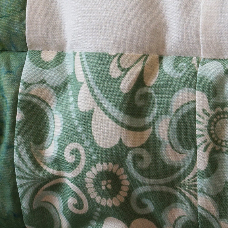 May include: Close-up of a quilt with a floral pattern in shades of green and white. The fabric features a repeating design of stylized flowers and swirling shapes, with white sections separating the patterned blocks.