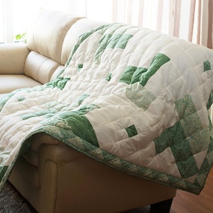 May include: A white and green quilt draped over a beige leather sofa. The quilt features a patchwork design with green geometric shapes and a green border. The sofa is in a well-lit room with a window and curtains.
