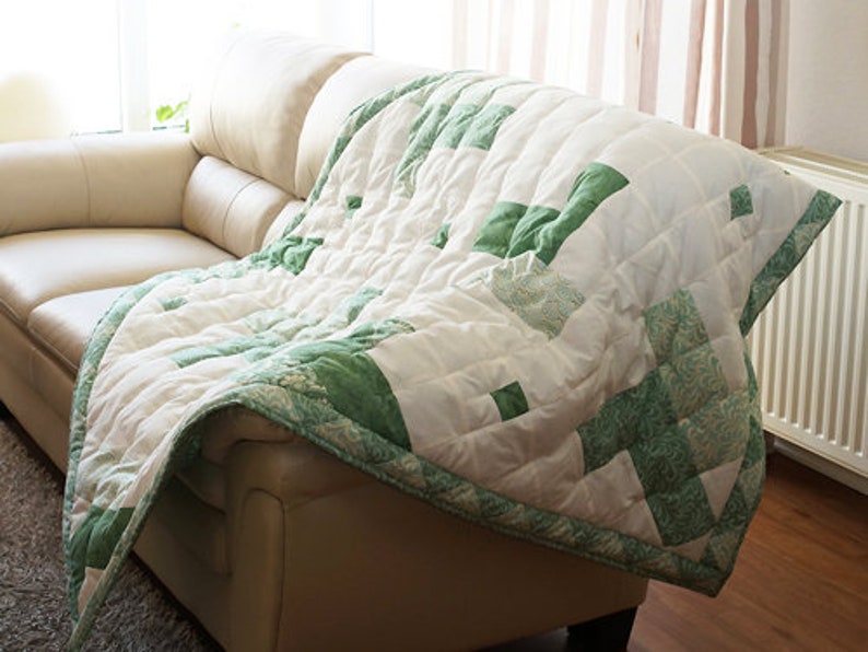 May include: A white and green quilt draped over a beige leather sofa. The quilt features a patchwork design with green geometric shapes and a green border. The sofa is in a well-lit room with a window and curtains.