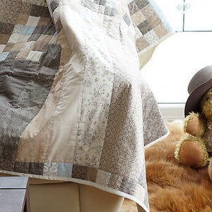 Pode incluir: Uma colcha de retalhos em tons de castanho, bege e azul claro, drapeada sobre uma cadeira. A colcha apresenta v&aacute;rios padr&otilde;es e texturas. Um urso de pel&uacute;cia com chap&eacute;u est&aacute; por perto. A cena evoca uma est&eacute;tica acolhedora e artesanal.