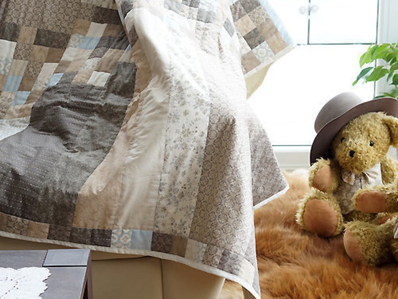 Pode incluir: Uma colcha de retalhos em tons de castanho, bege e azul claro, drapeada sobre uma cadeira. A colcha apresenta v&aacute;rios padr&otilde;es e texturas. Um urso de pel&uacute;cia com chap&eacute;u est&aacute; por perto. A cena evoca uma est&eacute;tica acolhedora e artesanal.