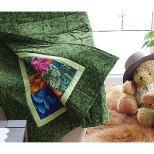 May include: A green quilt with a floral pattern and a contrasting floral square. The quilt is draped over a couch. The floral square features colorful flowers and fern patterns. The quilt is a cozy home decor item.