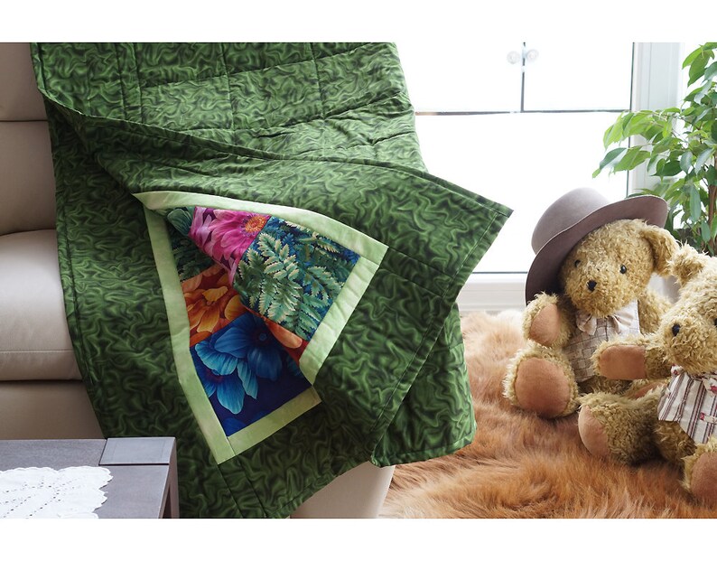 May include: A green quilt with a floral pattern and a contrasting floral square. The quilt is draped over a couch. The floral square features colorful flowers and fern patterns. The quilt is a cozy home decor item.
