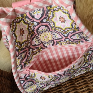 Puede incluir: Bolso de mano de tela con estampado de cuadros rosas y blancos y un estampado floral decorativo. El bolso tiene un bolsillo con cremallera y correas rosas. El estampado floral incluye elementos rosas, amarillos y marrones.