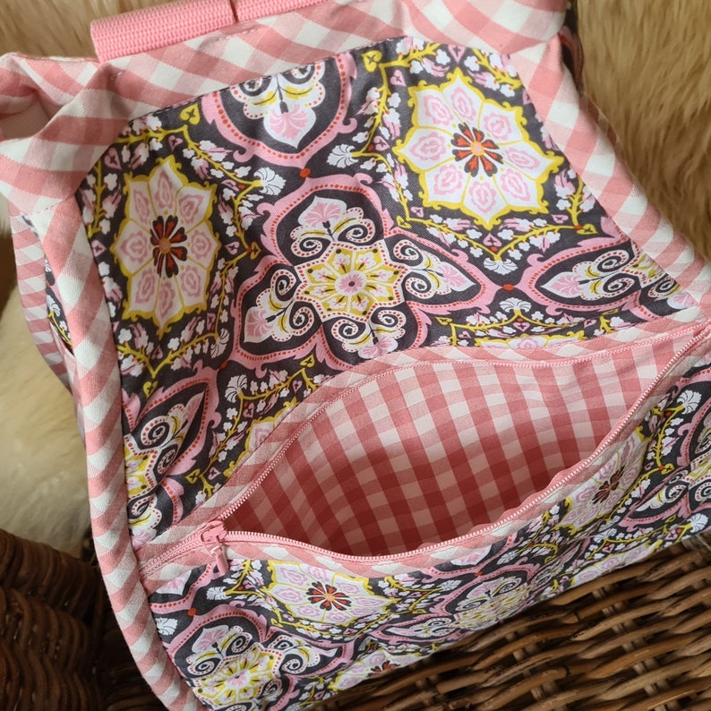 Puede incluir: Bolso de mano de tela con estampado de cuadros rosas y blancos y un estampado floral decorativo. El bolso tiene un bolsillo con cremallera y correas rosas. El estampado floral incluye elementos rosas, amarillos y marrones.