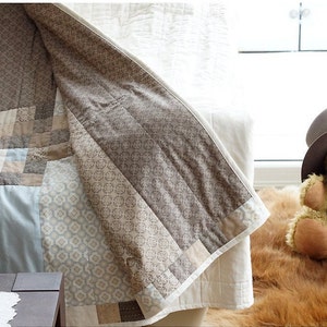 Pode incluir: Um edredom feito &agrave; m&atilde;o com um design de patchwork em tons de bege, castanho e azul claro. O edredom est&aacute; drapeado sobre um sof&aacute;, mostrando seus intrincados padr&otilde;es e texturas. Um urso de pel&uacute;cia com um chap&eacute;u est&aacute; por perto.