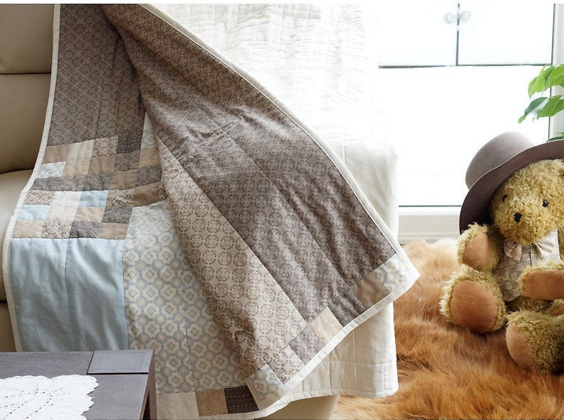 Pode incluir: Um edredom feito &agrave; m&atilde;o com um design de patchwork em tons de bege, castanho e azul claro. O edredom est&aacute; drapeado sobre um sof&aacute;, mostrando seus intrincados padr&otilde;es e texturas. Um urso de pel&uacute;cia com um chap&eacute;u est&aacute; por perto.