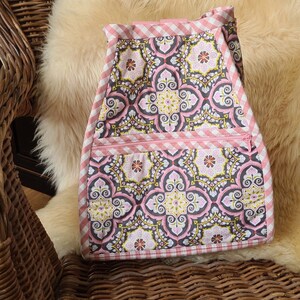 Puede incluir: Delantal de tela con estampado floral en rosa, gris y amarillo. El delantal tiene un ribete de cuadros rosas y un bolsillo con cremallera. El delantal est&aacute; sobre una superficie suave y clara.