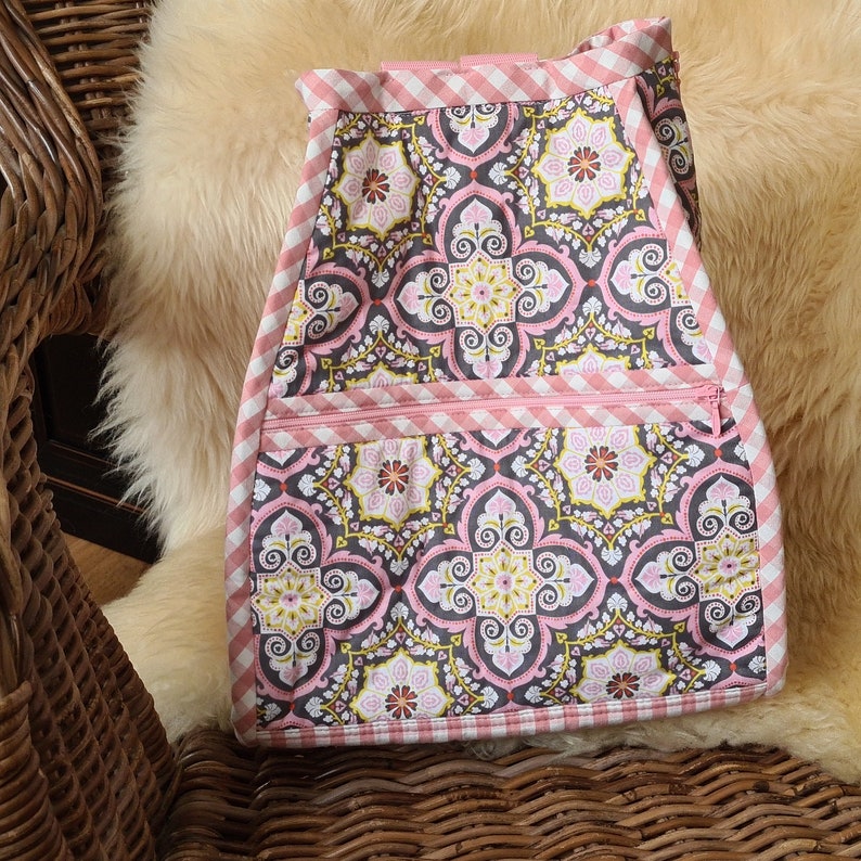 Puede incluir: Delantal de tela con estampado floral en rosa, gris y amarillo. El delantal tiene un ribete de cuadros rosas y un bolsillo con cremallera. El delantal est&aacute; sobre una superficie suave y clara.