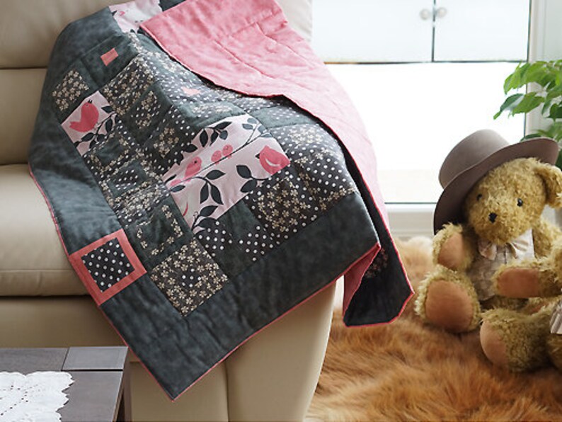 May include: A handmade quilt with a patchwork design featuring gray, pink, and white fabrics. The quilt has a floral and polka dot pattern, with a pink border. A teddy bear wearing a hat sits nearby.