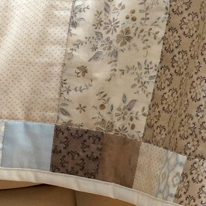 Pode incluir: Um edredom de patchwork com v&aacute;rios tecidos estampados em tons de bege, castanho e azul. O edredom incorpora desenhos florais, pontilhados e geom&eacute;tricos, criando uma est&eacute;tica vintage. As bordas s&atilde;o finalizadas com uma borda branca.