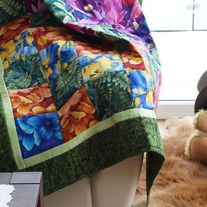 May include: A colorful floral quilt with a green border. The quilt features squares of blue, orange, and yellow flowers and green foliage. The quilt is draped over a white surface. A teddy bear wearing a hat sits nearby.