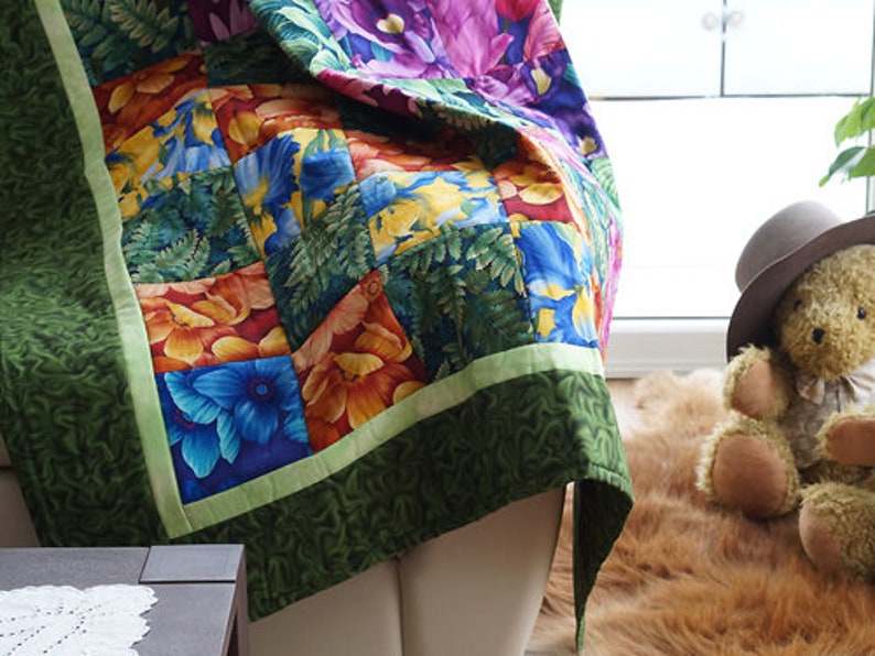 May include: A colorful floral quilt with a green border. The quilt features squares of blue, orange, and yellow flowers and green foliage. The quilt is draped over a white surface. A teddy bear wearing a hat sits nearby.