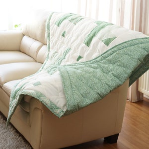 May include: A white and green quilt draped over a cream-colored sofa. The quilt features a geometric pattern with green accents and a patterned green border. The sofa is in a well-lit room with a rug.