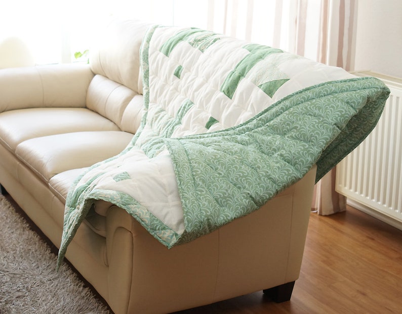 May include: A white and green quilt draped over a cream-colored sofa. The quilt features a geometric pattern with green accents and a patterned green border. The sofa is in a well-lit room with a rug.