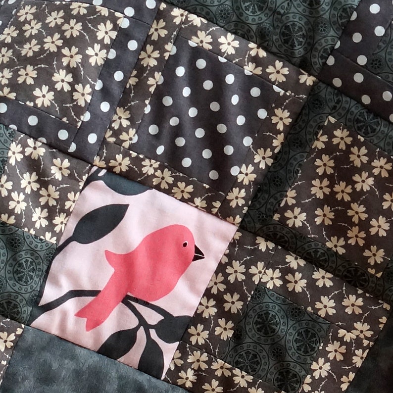 May include: Close-up of a patchwork quilt with various fabric patterns. The quilt features squares with floral, polka dot, and bird designs in shades of brown, gray, and pink. The stitching is visible, highlighting the quilt's construction.