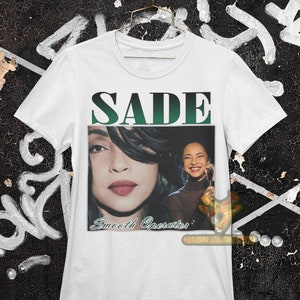 May include: White t-shirt with a green and black graphic of Sade with the text "SADE" and "Smooth Operator" printed on the front.