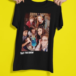 Puede incluir: Camiseta negra con un estampado blanco del reparto de That 70s Show. El estampado muestra a los personajes en un ambiente de sala de estar, con un líquido azul en un recipiente de vidrio en el centro.