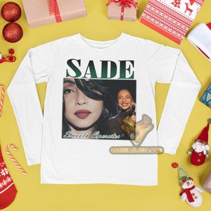 May include: White long-sleeve shirt with a graphic print of Sade, a singer, with the text "SADE" and "Sweetest Operator" on the front.